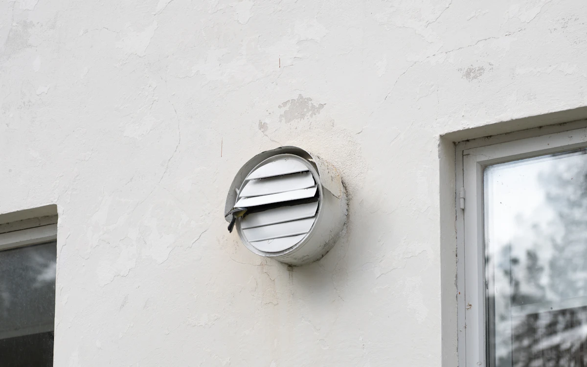 Grille d'aération ronde à lamelles sur une façade blanche, à côté d'une fenêtre